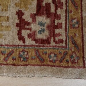 Close-up of a rug with beige, red, and gold geometric motifs, a textured pile, and visible edge fringe.