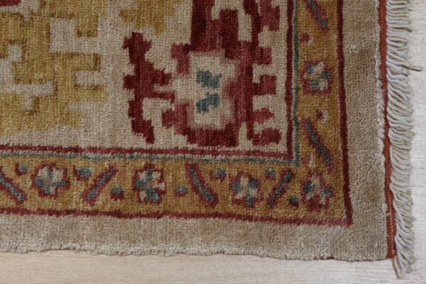 Close-up of a rug with beige, red, and gold geometric motifs, a textured pile, and visible edge fringe.