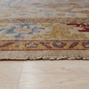 The textured edge of a light-colored rug featuring gold, rust, and blue patterned borders on wood flooring.