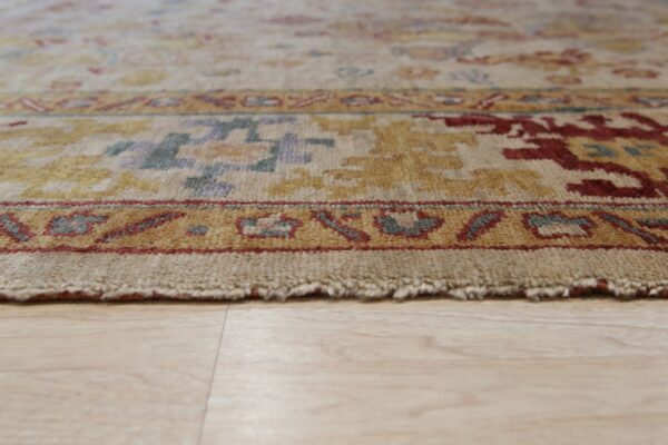 The textured edge of a light-colored rug featuring gold, rust, and blue patterned borders on wood flooring.