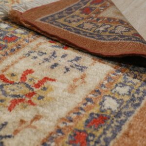 A patterned beige, blue, and rust high-pile rug is folded back, showing its woven border on wood floor.