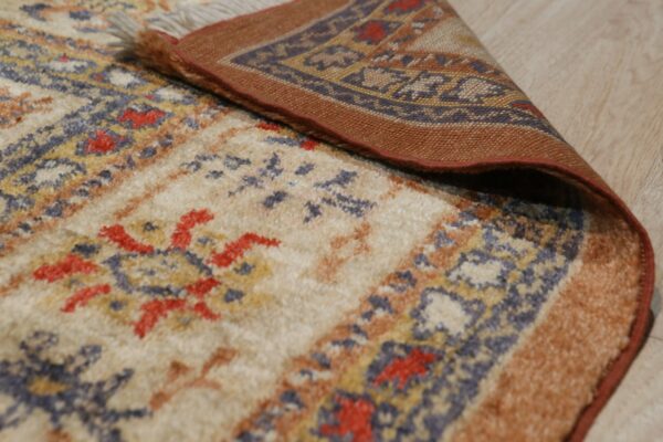 A patterned beige, blue, and rust high-pile rug is folded back, showing its woven border on wood floor.