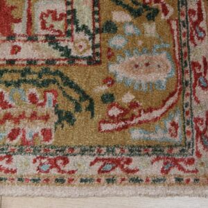 Traditional rug corner with fringe, featuring gold, rust, green, and pale blue patterns on wood flooring.