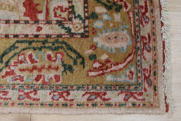 Traditional rug corner with fringe, featuring gold, rust, green, and pale blue patterns on wood flooring.