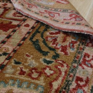 Textured rug detail showing red, brown, and blue traditional patterns, folded over on light wood flooring.