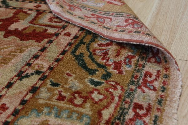 Textured rug detail showing red, brown, and blue traditional patterns, folded over on light wood flooring.