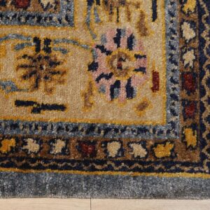 Tan rug corner with blue borders and intricate traditional patterns in gold, navy, and pink on wood floor.
