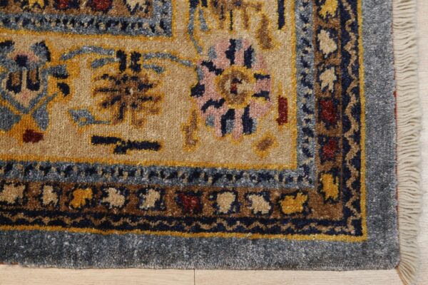 Tan rug corner with blue borders and intricate traditional patterns in gold, navy, and pink on wood floor.