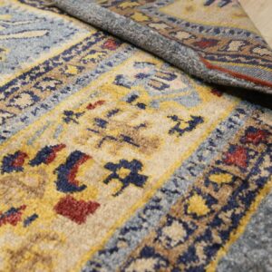 Close-up shows a medium pile blue, yellow, and beige patterned rug curled on light wood flooring.