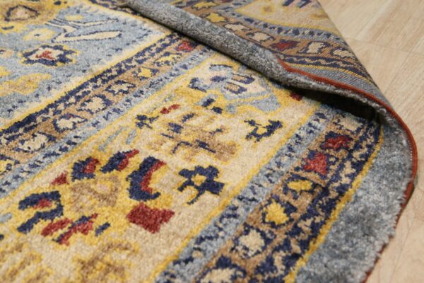 Close-up shows a medium pile blue, yellow, and beige patterned rug curled on light wood flooring.