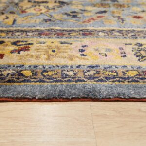Low-angle detail of a blue, gold, and red patterned rug edge on light blonde wood flooring.