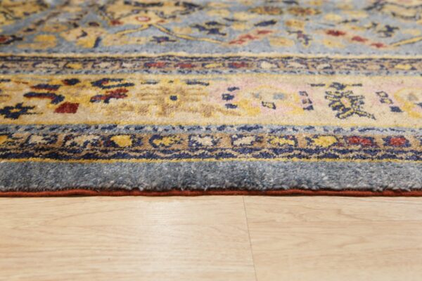 Low-angle detail of a blue, gold, and red patterned rug edge on light blonde wood flooring.