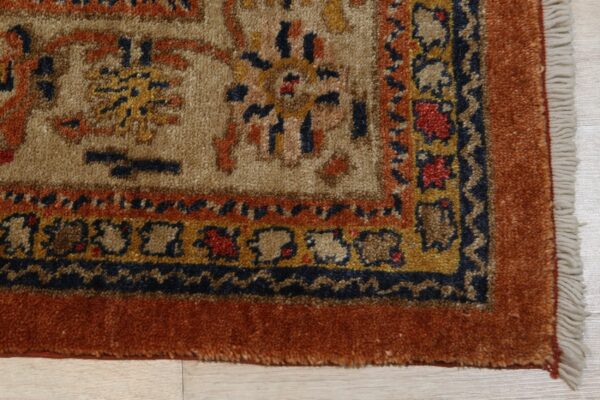 Traditional thick-pile rug corner featuring tan field, rust border, and blue and gold accents on wood.