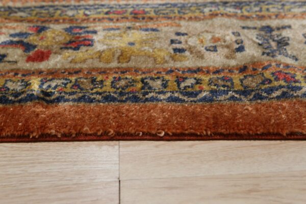 Textured patterned rug in beige, blue, and orange colors with a thick border resting on light wood flooring.
