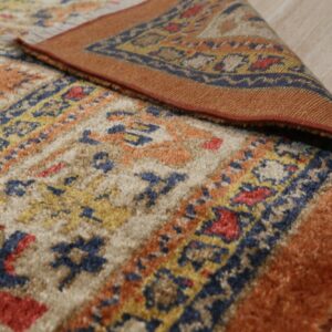 A textured rug in rust, navy, and cream tribal patterns is curled back on light flooring.