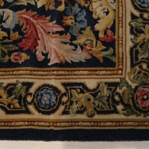 Corner detail of a medium pile rug featuring navy, gold, red, and green floral patterns within a tan border.