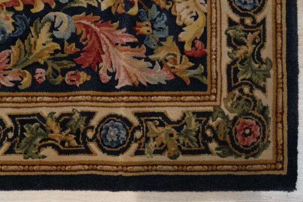 Corner detail of a medium pile rug featuring navy, gold, red, and green floral patterns within a tan border.