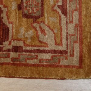 Textured rug corner displaying geometric patterns in gold, red, and beige, resting on wood flooring.