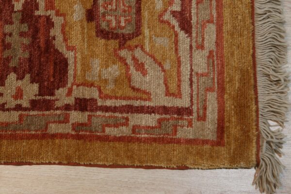 Textured rug corner displaying geometric patterns in gold, red, and beige, resting on wood flooring.