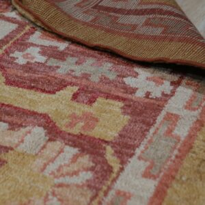 A rust, yellow, and beige geometric patterned rug is folded back to reveal the woven backing on wood flooring.