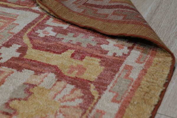 A rust, yellow, and beige geometric patterned rug is folded back to reveal the woven backing on wood flooring.