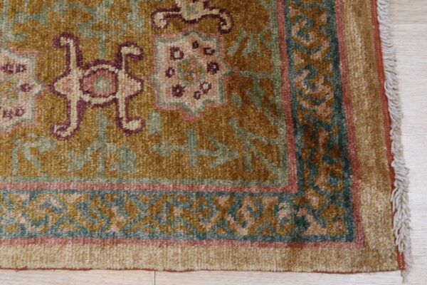Textured golden rug detail with traditional patterns, burgundy accents, teal border, and light fringe.