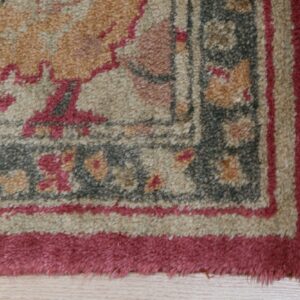 Textured rug corner displaying ornate patterns in beige, pink, and grey, with a visible fringe on light wood.
