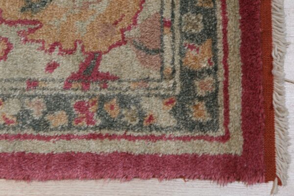 Textured rug corner displaying ornate patterns in beige, pink, and grey, with a visible fringe on light wood.