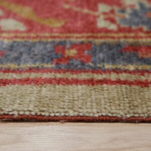 Textured close-up of a thick pile rug edge showing beige, red, and slate blue patterns on light wood flooring.