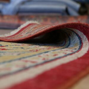 Close up of a yellow, red, and blue patterned rug with a red border, curled to reveal its short pile and fringe.