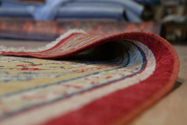 Close up of a yellow, red, and blue patterned rug with a red border, curled to reveal its short pile and fringe.