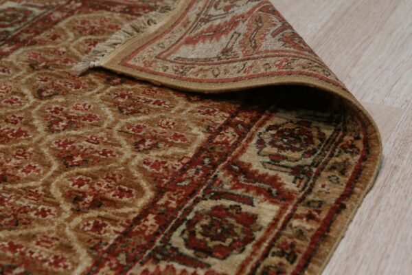 A tan and rust traditional rug with a low pile is curled back on light wood flooring.