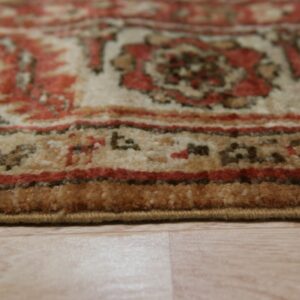Close-up of a medium-pile rug displaying traditional red, cream, and tan patterns on light wood flooring.