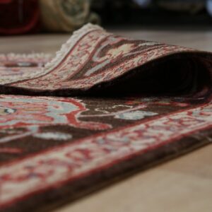 A traditional patterned rug with brown, red, and blue tones is curled back on a light wood floor.
