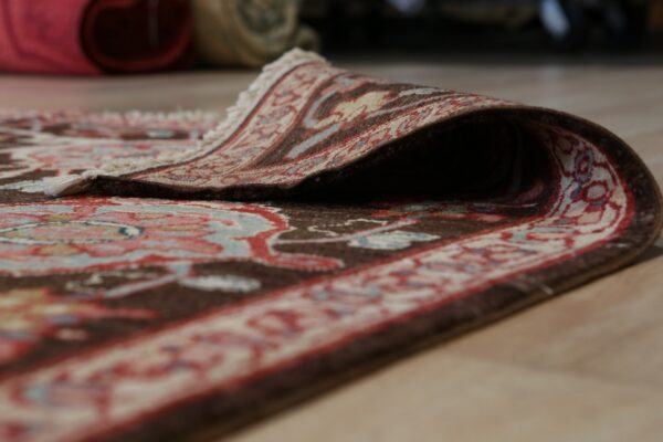 A traditional patterned rug with brown, red, and blue tones is curled back on a light wood floor.