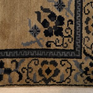 Tan pile rug with black and slate gray floral and geometric border detail on light wood floor.