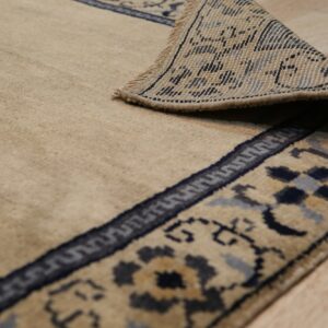 The corner of a tan rug with a detailed blue and beige border is curled back on wood flooring.