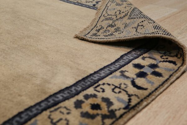 The corner of a tan rug with a detailed blue and beige border is curled back on wood flooring.