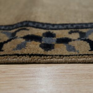 Close view of a tan, medium-pile rug edge with black and gray border motifs on light wood planks.