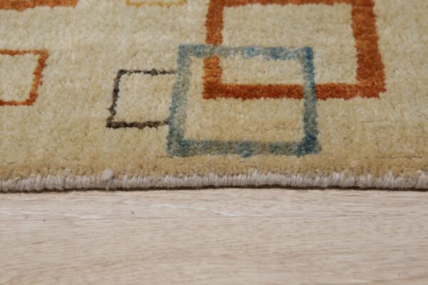 Beige medium pile rug with blue, rust, and brown geometric squares rests on light wood grain floor.