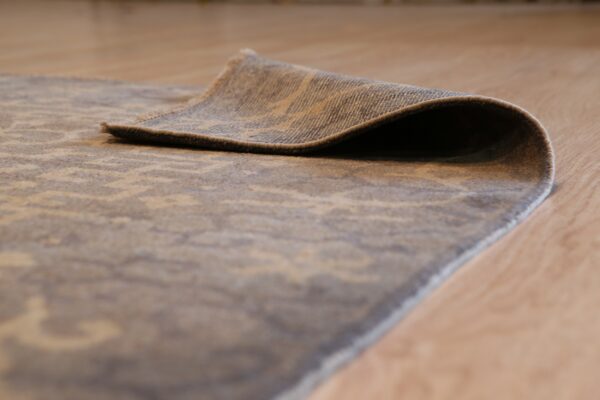 Muted beige and grey patterned rug edge is curled up on a light hardwood floor.