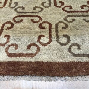 Textured beige rug featuring brown scrolling designs, a dark border, and light fringe on wood planks.