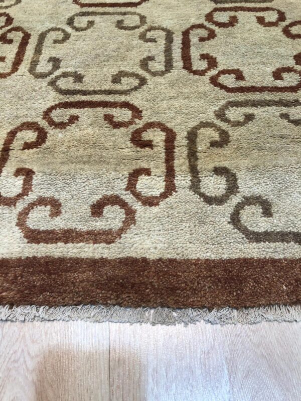 Textured beige rug featuring brown scrolling designs, a dark border, and light fringe on wood planks.