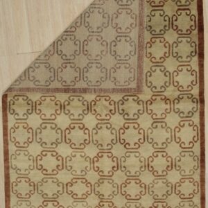 Tan rug with repeating brown geometric scroll pattern and border rests on light wood flooring.