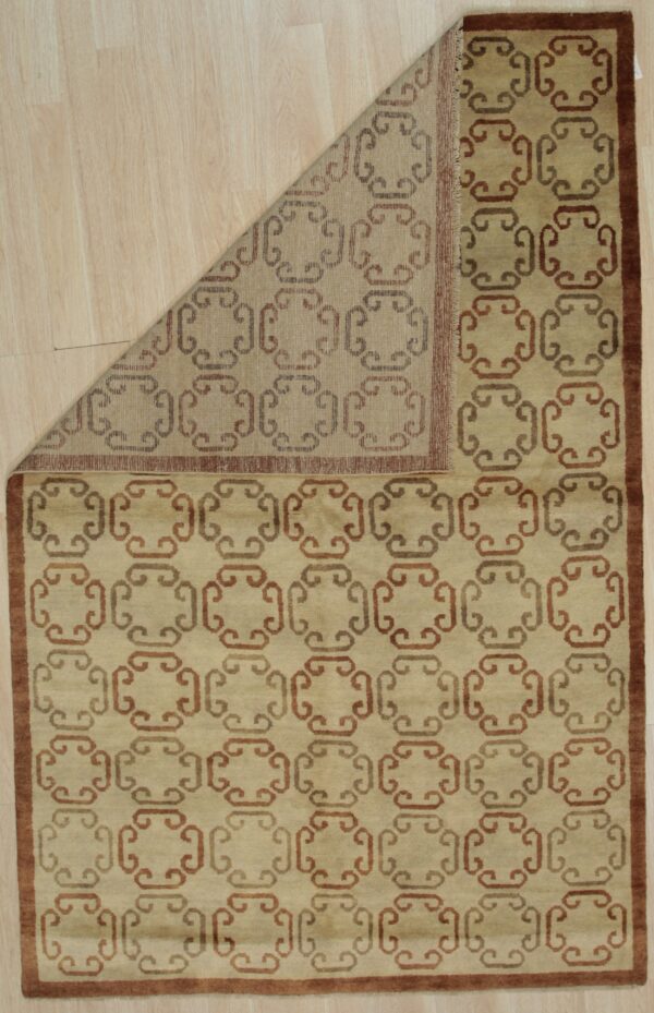 Tan rug with repeating brown geometric scroll pattern and border rests on light wood flooring.
