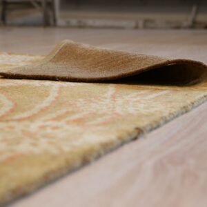 A beige rug with subtle coral patterns is lifted on light wood flooring, showing the brown backing.
