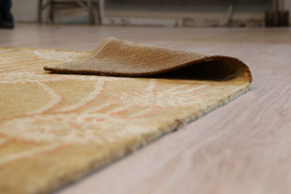 A beige rug with subtle coral patterns is lifted on light wood flooring, showing the brown backing.