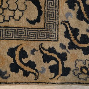 Tan rug detail featuring a black Greek key border and dark floral motifs on light wood.