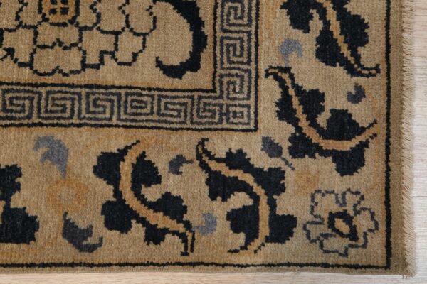 Tan rug detail featuring a black greek key border and dark floral motifs on light wood.