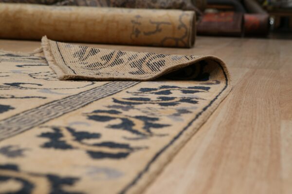 Cream and blue patterned rug partially unrolled on a wood floor, showing the low pile and woven backing.